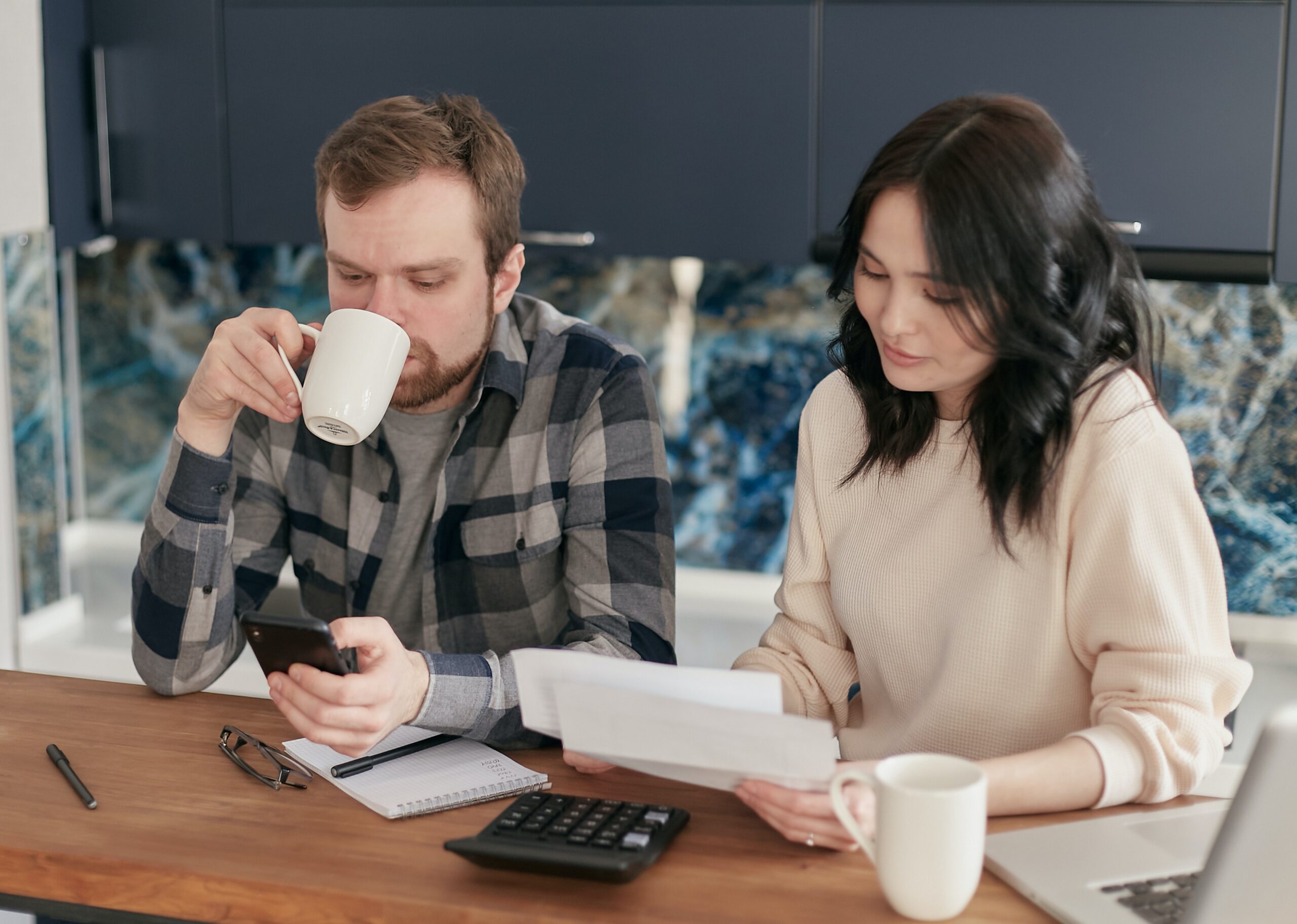man sipping coffee scrolling his phone sitting next to woman reading papers