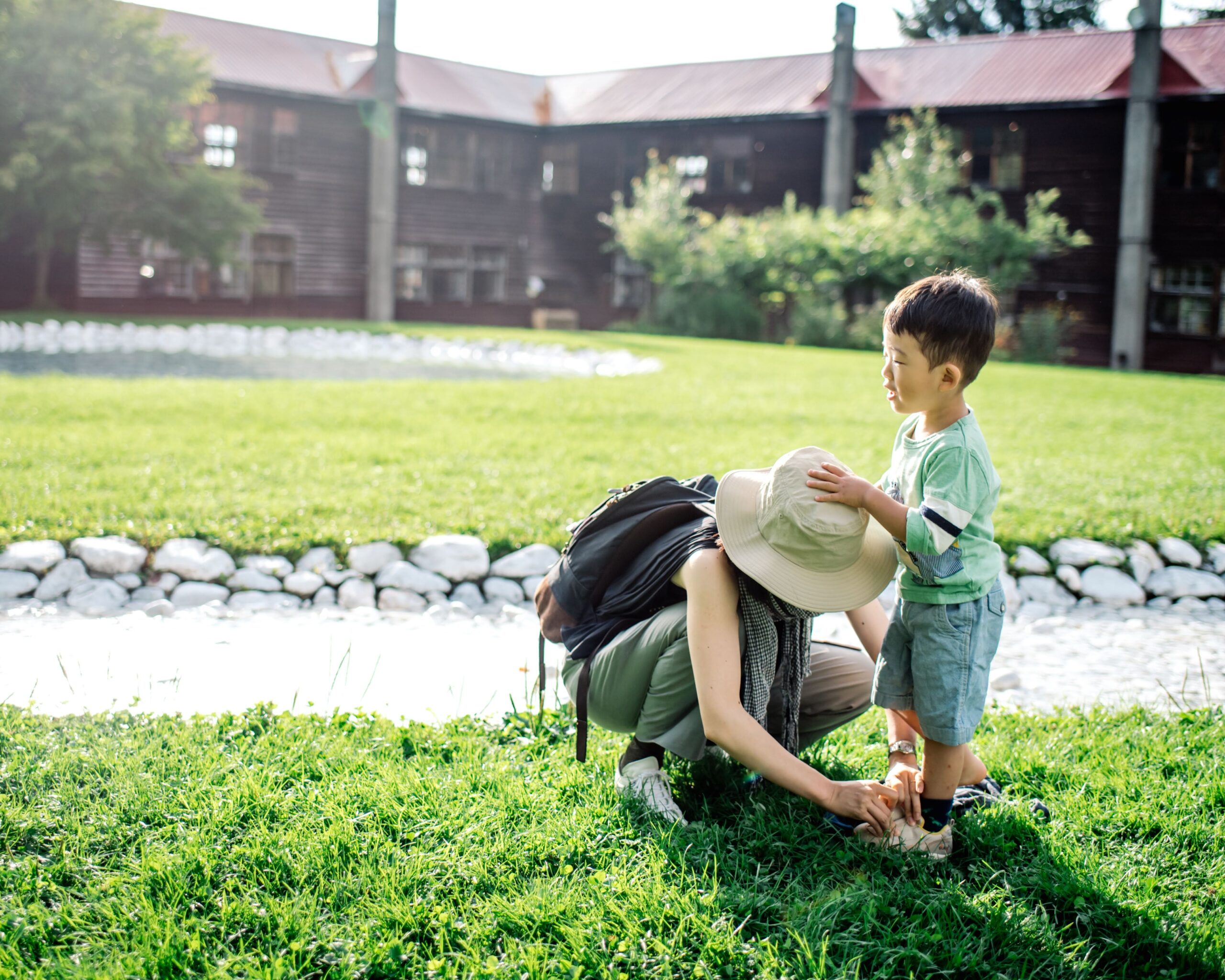 mother bent over tying her child's shoe wearing a big floppy hat outside.