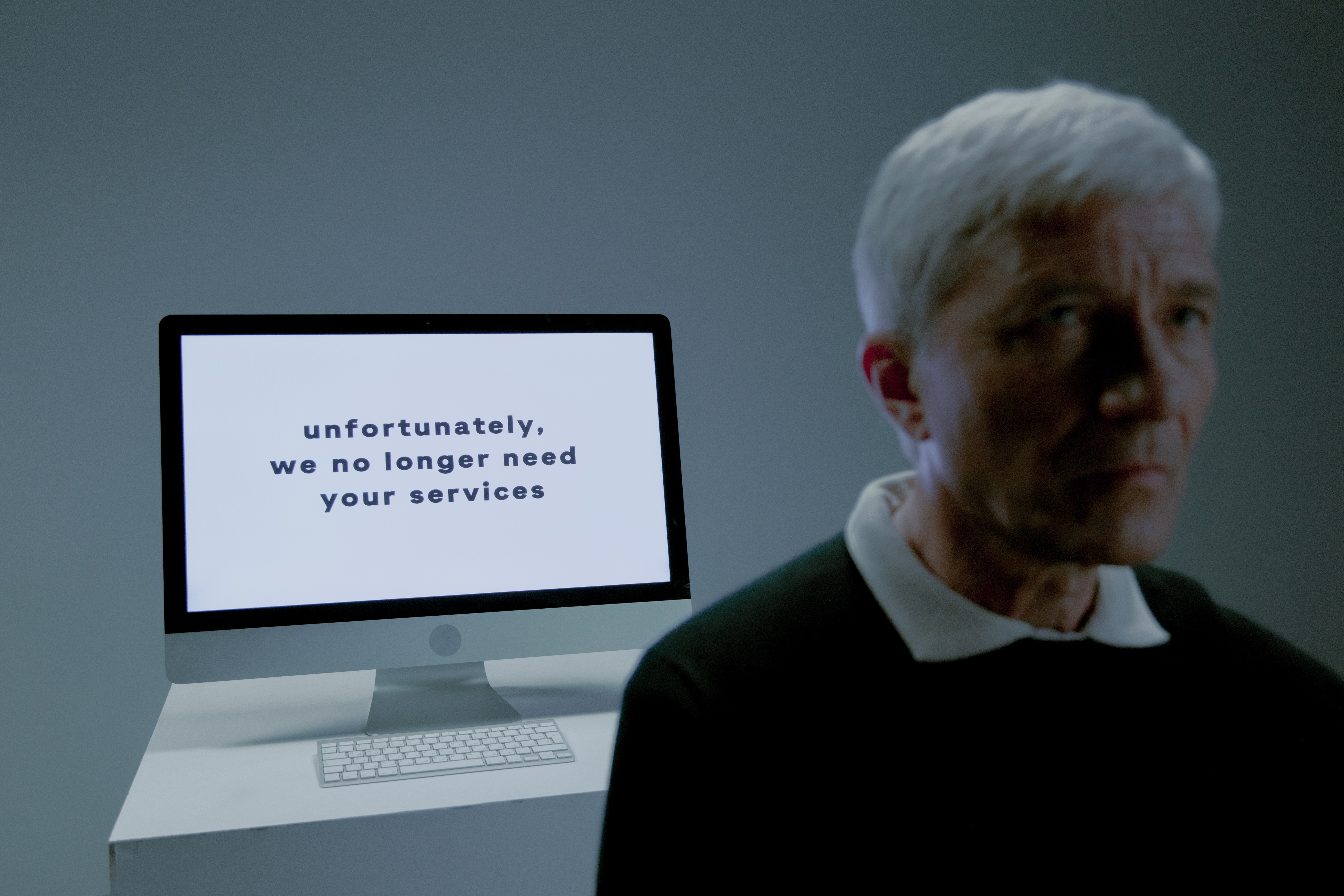 older man in front of computer with "unfortunately we no longer need your services" on the screen