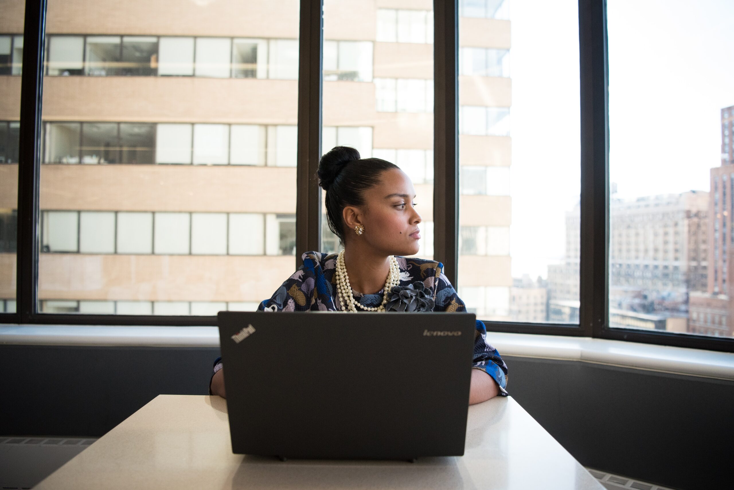 woman with brown skin sitting at a table in front of a laptop and looking out the window