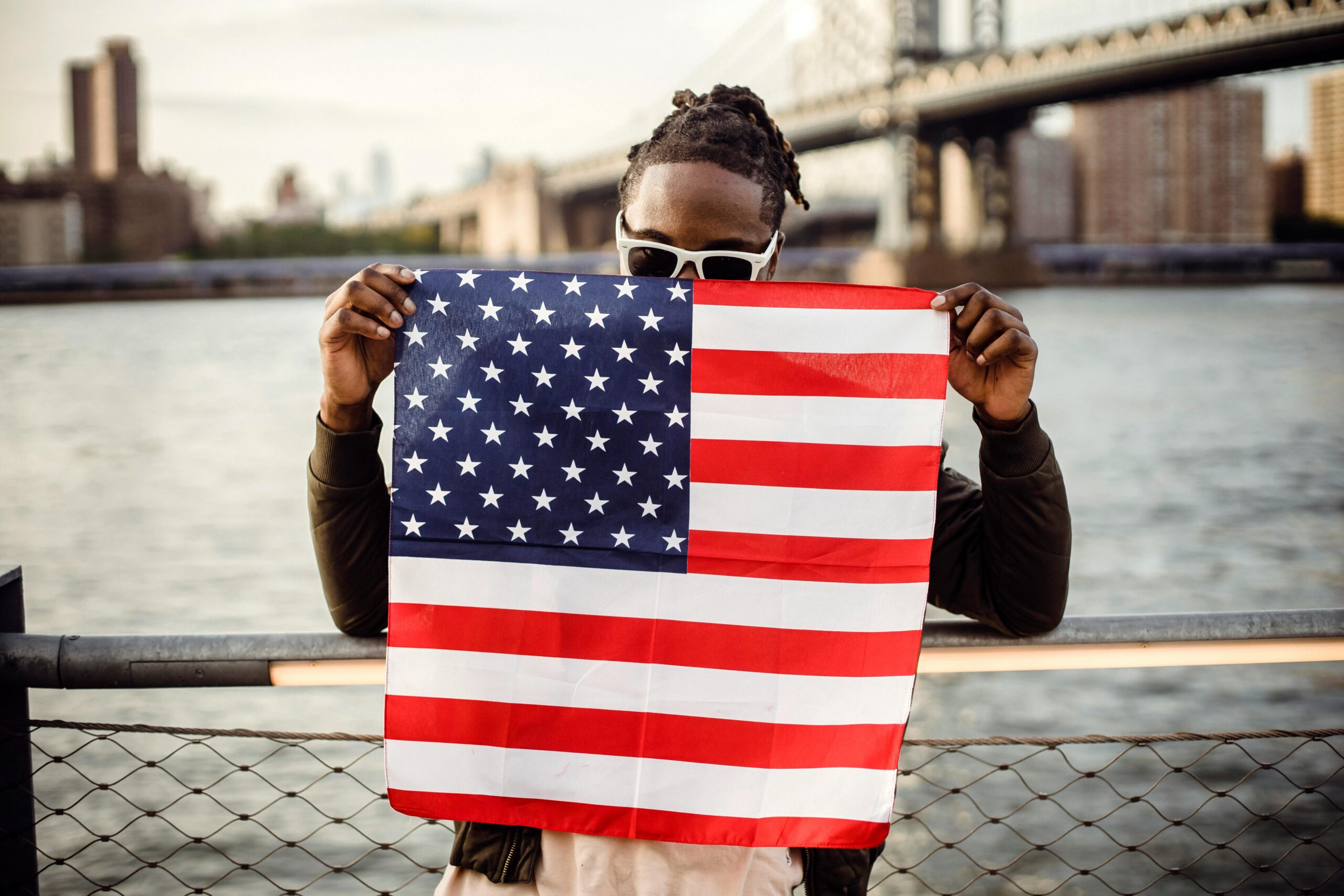 black man wearing sunglasses holding American flag in front of his face