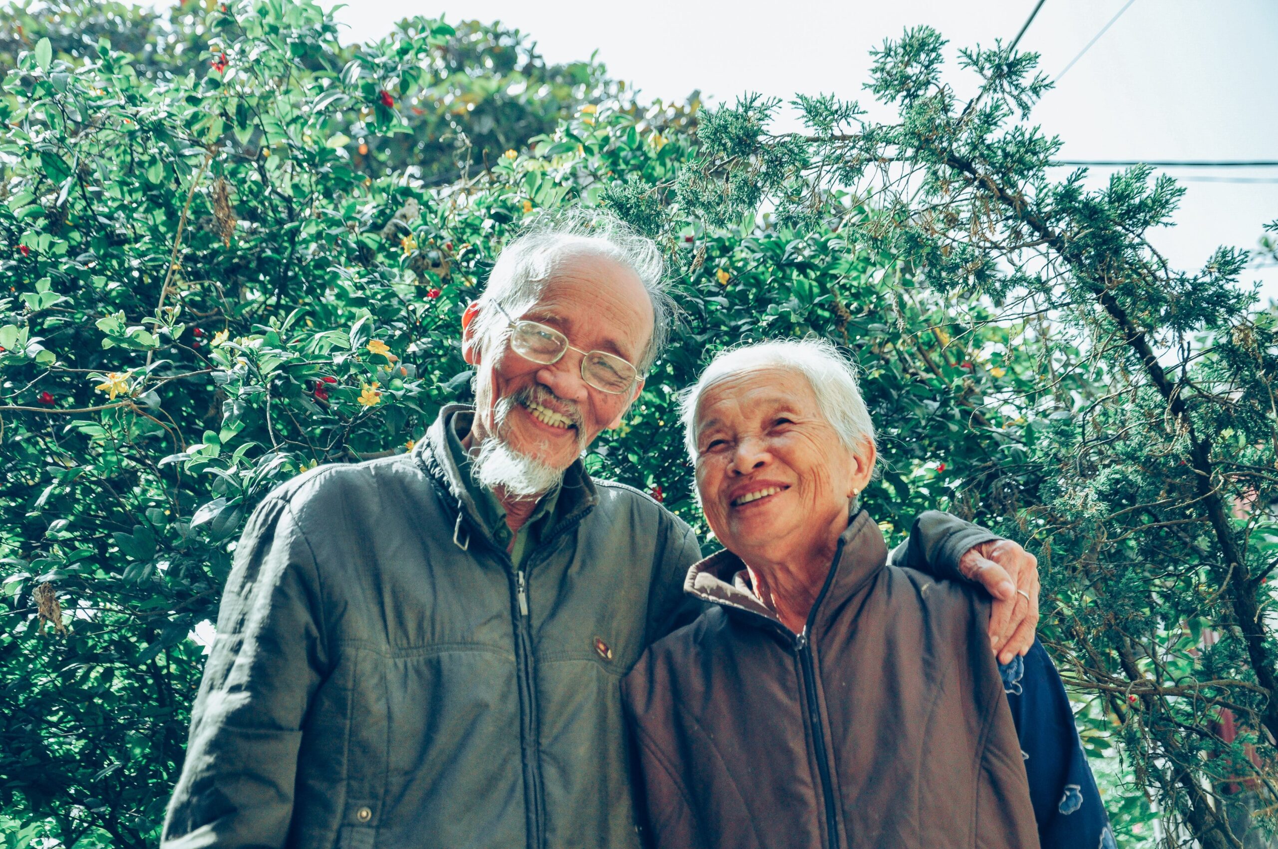 older man with arm around smiling older woman standing in front of trees