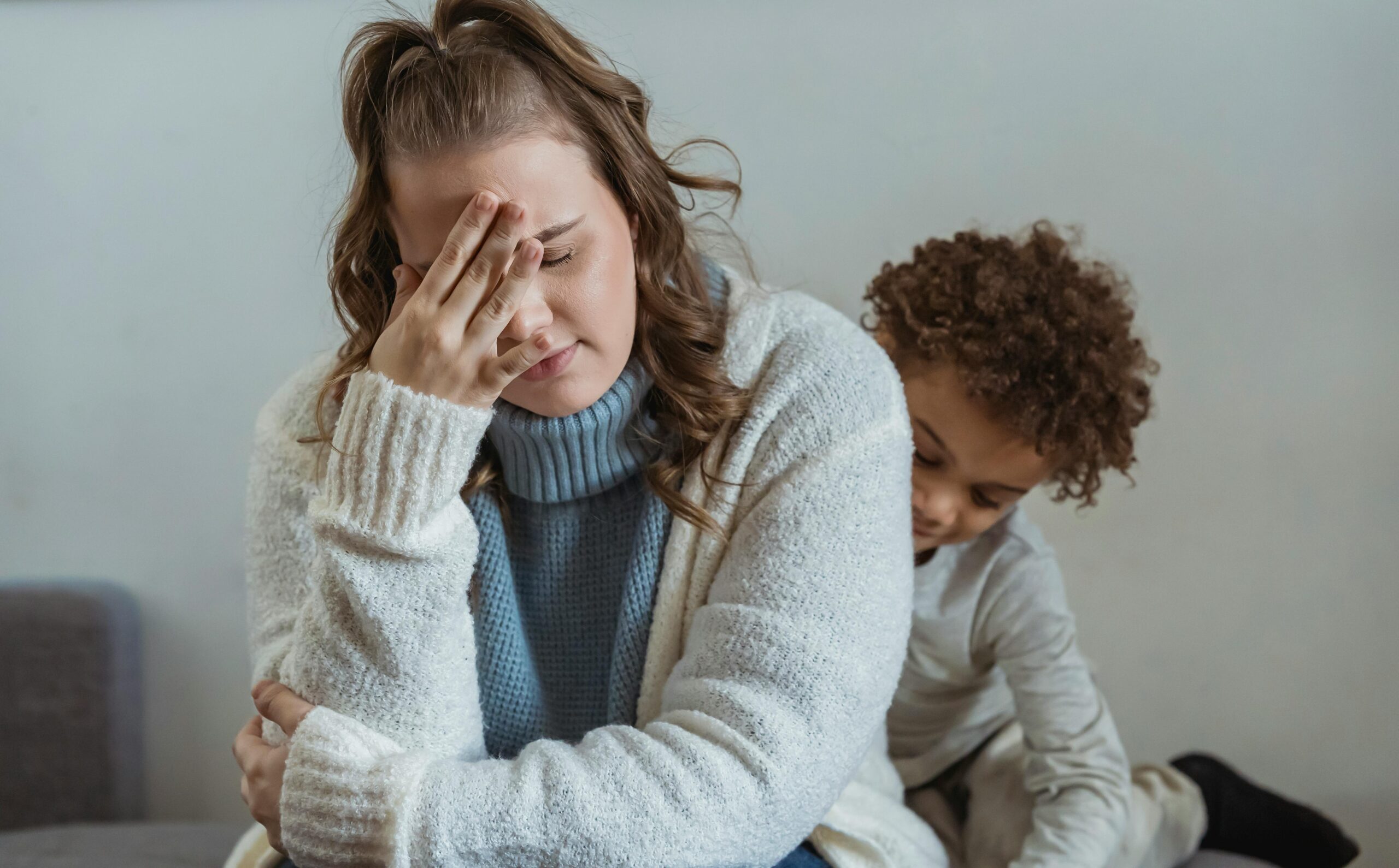 child peeks out from behind a tired mother with face in hand